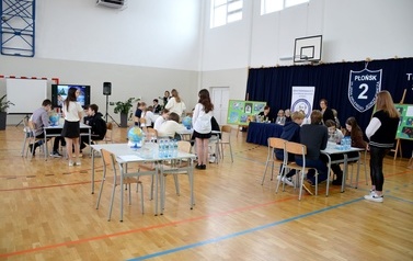 grupy os&oacute;b siedzą przy stołach w sali gimnastycznej