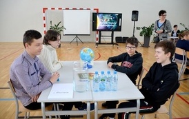 grupa os&oacute;b siedzi przy stole na kt&oacute;rym jest globus oraz butelki z wodą